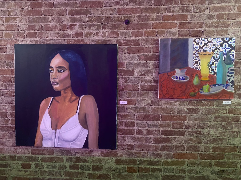 Two paintings, the one on the left of a Somalian woman looking off to the left and wearing a white tank top. The painting on the left has a dark background. The painting on the right is a still life utilizing an exaggerated style with brighter colors.